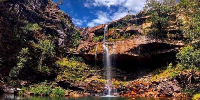 Waterfall cascading down a rocky cliff into a shallow pool, surrounded by green trees and people exploring the rocks below.
