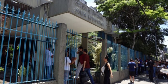 People line up outside a concrete building with a bright blue spiked fence and surrounding greenery under a sunny sky while waiting to enter.