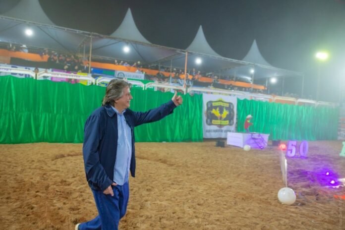Man in a blue jacket walking on a sandy arena at night, giving a thumbs-up with illuminated event stands in the background.