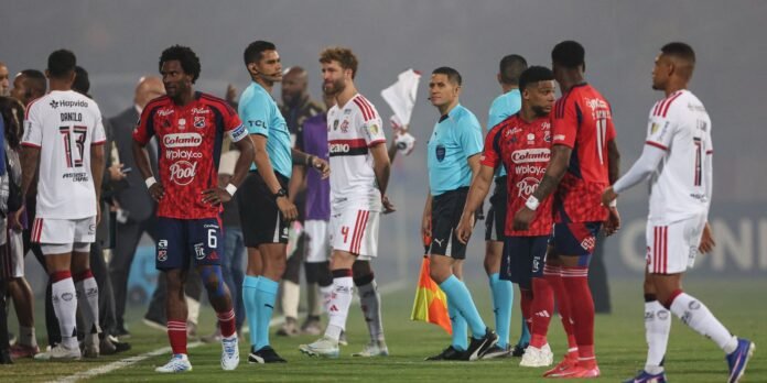 Football players from two teams line up on the pitch after a match, with referees in turquoise shirts nearby.