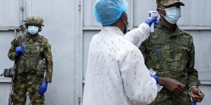 Healthcare worker in a white protective gown and blue cap checks a soldier's temperature with an infrared thermometer; both wear masks and gloves, with another soldier in camouflage in the background.