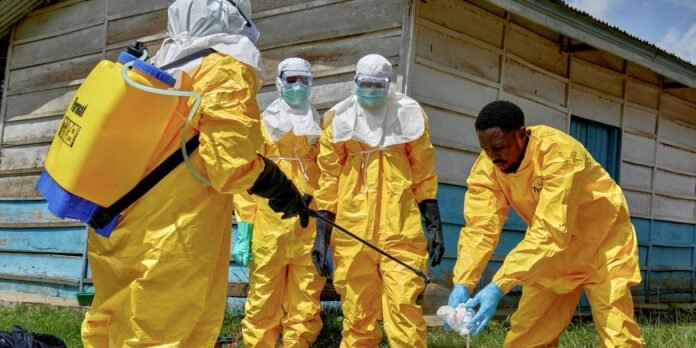 Team of four people in yellow hazmat suits spraying disinfectant outside a weathered wooden building, wearing masks and gloves.