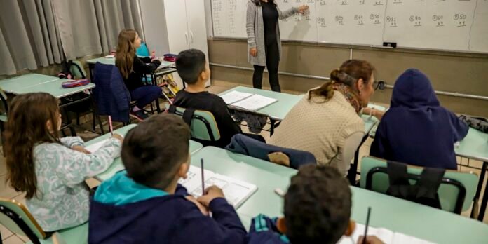 Teacher writes math work on whiteboard as students take notes in a classroom.