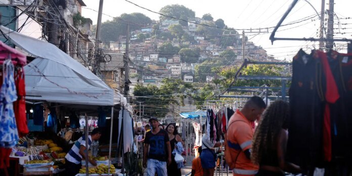 Bustling outdoor market along a street with fruit stalls and clothing racks, hillside homes in the background.