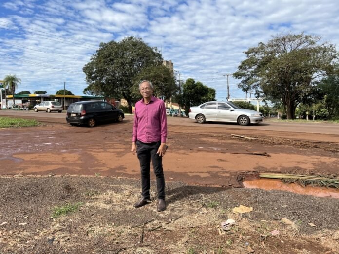 Man in a magenta shirt standing on a dusty street with parked cars and trees behind him.