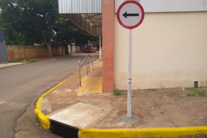 Left-turn traffic sign mounted on a pole at a sidewalk next to a building, with a yellow curb and a small ramped sidewalk area nearby.