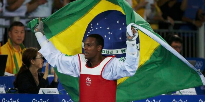 Brazilian flag raised by a taekwondo athlete in a white gi with red chest guard, celebrating at Rio 2016.