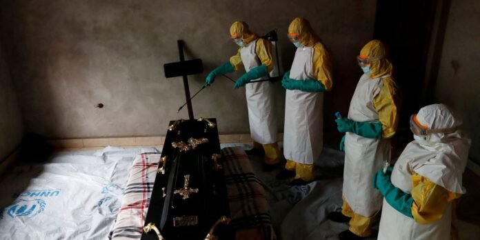 Four people in full PPE (yellow suits, green gloves, masks) stand around a black coffin in a dim room, one holding a spraying wand over the coffin edge.