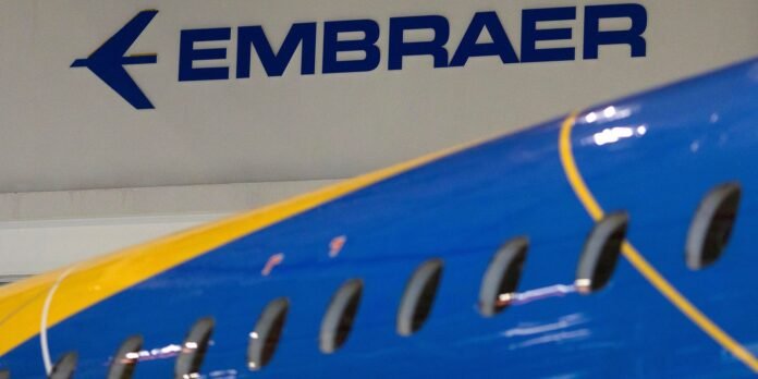 Blue and yellow Embraer aircraft fuselage in a hangar; Embraer logo on wall in background.