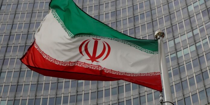 Iranian flag waving on a flagpole against a glass-fronted office building, showing green, white, and red stripes with the emblem in the center and Kufic borders along the white stripe.