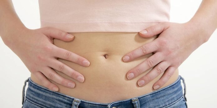 Close-up of a person's bare abdomen with hands resting on either side of the navel, wearing jeans and a light pink top.