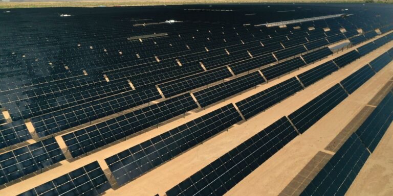 A vast field of dark solar panels arranged in neat rows on a sandy desert landscape, reflecting sunlight.