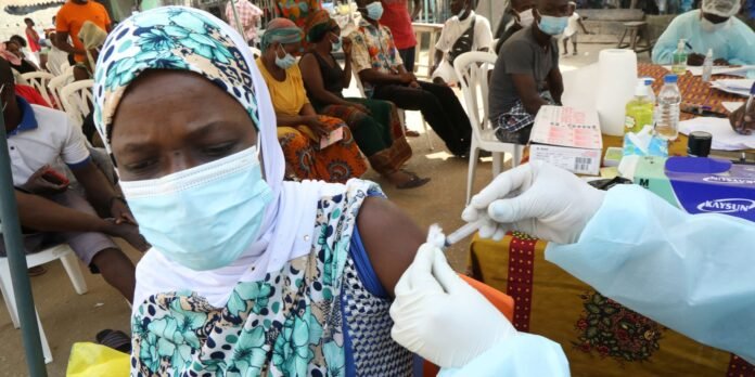 A woman wearing a patterned headscarf and mask receives a vaccination in her upper arm from a gloved healthcare worker at an outdoor clinic.