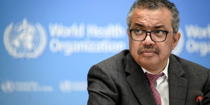 Man in a dark suit and red tie at a WHO press briefing, looking to the side on a blue backdrop.