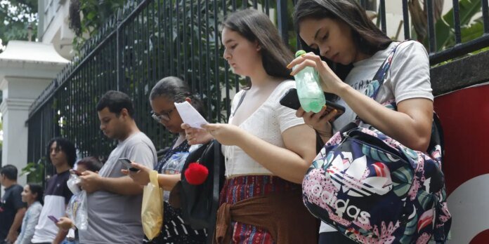 People line up along a black fence outdoors, each absorbed in a phone or papers, waiting patiently.
