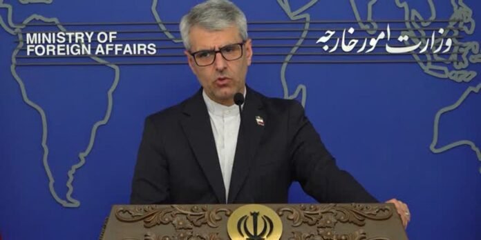 Spokesperson in a suit at a carved podium delivering a briefing against a blue backdrop marked Ministry of Foreign Affairs on the left and Persian script on the right.