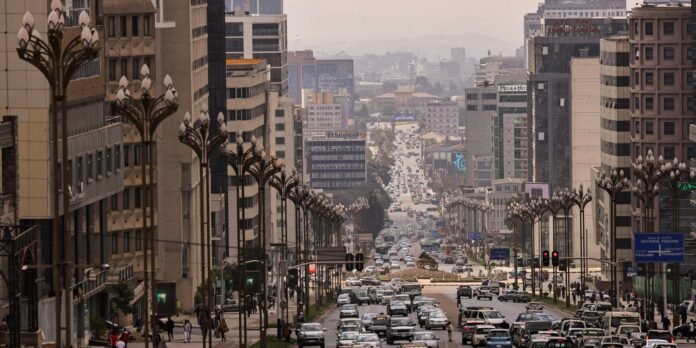 Wide urban avenue flanked by tall modern buildings and ornate street lamps, heavy traffic, and pedestrians along the sidewalks as the road recedes into the hazy distance.