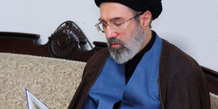 Bearded man with glasses and a black turban reads a document while wearing a blue vest and brown robe, seated on a patterned sofa.