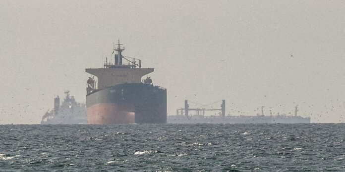 A large bulk carrier sails toward the camera on a choppy sea, with a distant port and cranes visible on the horizon and seabirds flying overhead.