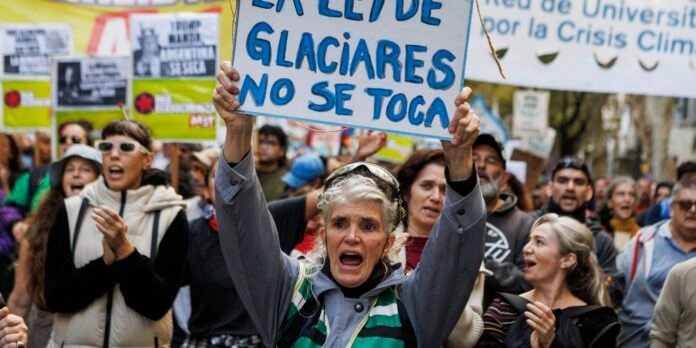 Protester shouting while raising a sign that reads 'LOS GLACIARES NO SE TOCA' at a climate march. Central woman in gray jacket, crowd behind her.