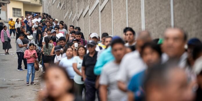 A long queue of diverse people standing on a street beside a tall concrete wall, waiting in line.