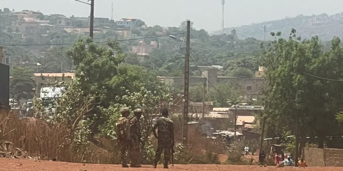 Two armed soldiers in camouflage stand on a dusty hill overlooking a small town with trees and utility poles in the background, during daytime.