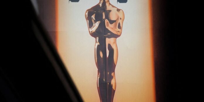 Golden Oscar-style statuette statue illuminated against dark background, standing upright with arms crossed.