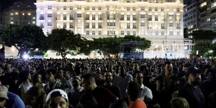 A large crowd of people gathered outdoors at night in front of a brightly lit, grand white building.
