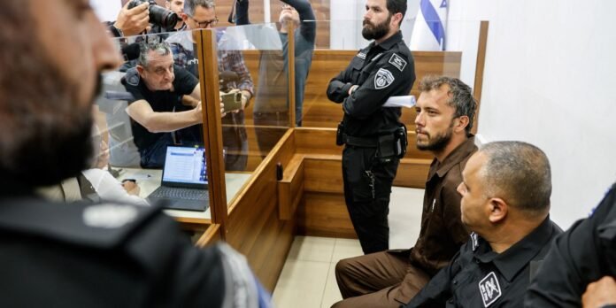Defendant in brown jail suit seated at the bench, guarded by two police officers, while journalists and a photographer observe through a glass partition.