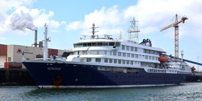 Large white-and-navy luxury yacht named Hondius docked at a harbor with shipyard buildings and a crane in the background.