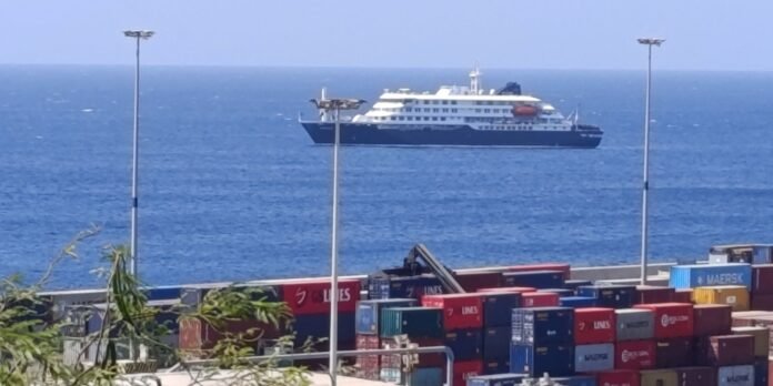 Port scene with stacked shipping containers in the foreground and a large white-and-navy ferry on calm blue sea in the distance,” with the ship near the horizon.