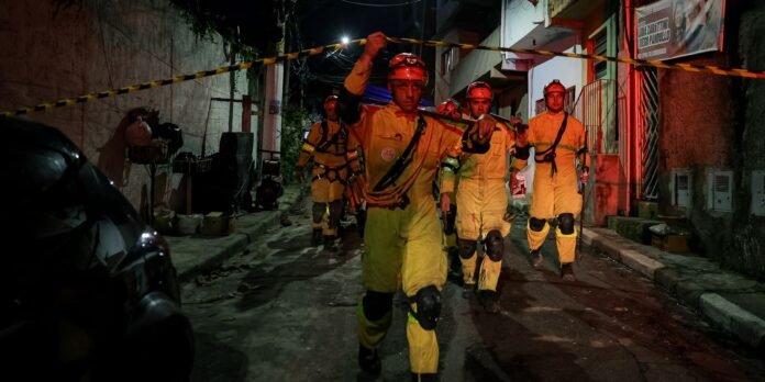 Firefighters in yellow protective suits march down a dark street behind yellow caution tape at night.