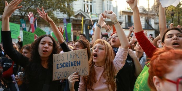 Crowd of young protesters on a city street, raising arms and shouting, a cardboard sign reads 'Los lápices escriben todos los días' in the foreground.