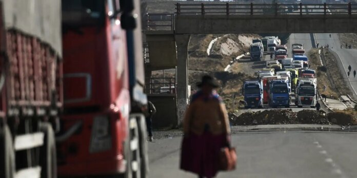 Queue of colorful trucks lined up on a highway beneath a concrete overpass, with a blurred pedestrian walking toward the camera in the foreground.