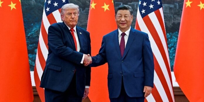 Two men in dark suits shake hands in front of US and Chinese flags.