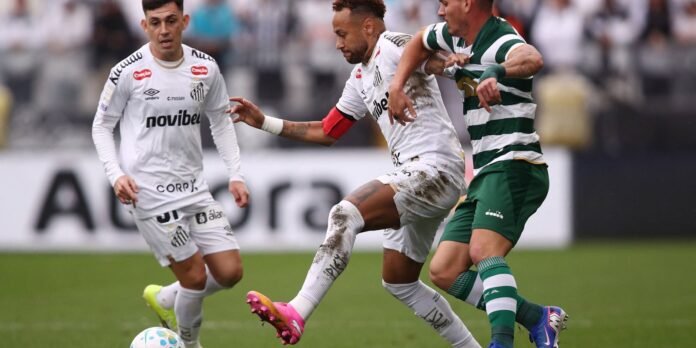 Three soccer players contest the ball: two in white kits and one in green-and-white stripes, on a muddy pitch with blurred stands.