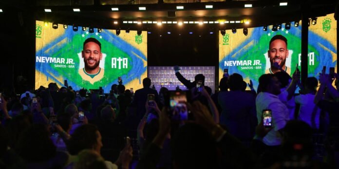 Stage with two large screens showing a smiling soccer player in a Brazil jersey; crowd records with phones at a stadium event.