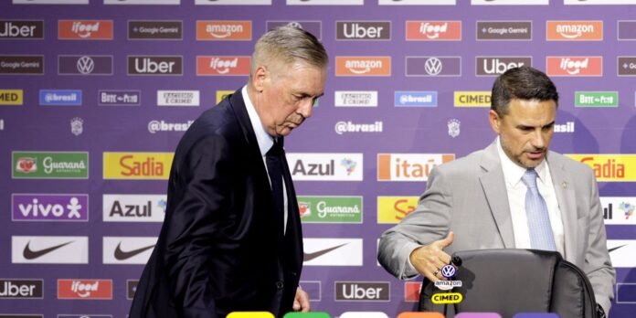 Two men in suits at a sponsorship-branded press conference backdrop; the man on the left looks down while the man on the right sits beside a chair.