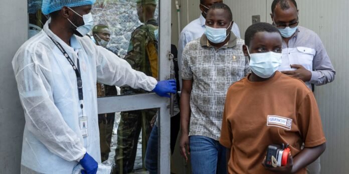 Healthcare worker in full PPE (gown, gloves, mask, cap) guiding a group of masked men through a doorway.