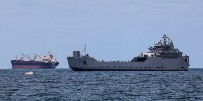 Large gray military vessel at sea with helicopter/flight deck and raised bridge, foreground; a red-and-white cargo ship in the distance to the left.