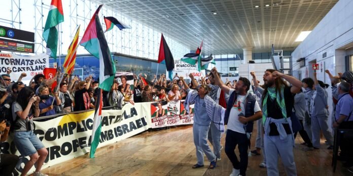 Crowd of protesters at an airport arrivals hall waving Palestinian flags and holding banners and posters while photographers document the scene.
