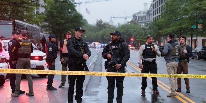Police officers in tactical gear stand behind yellow 'Police Line Do Not Cross' tape blocking a city street.