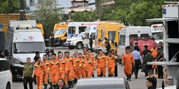 Group of emergency responders in orange jumpsuits walking in a lane lined with vans and ambulances, with officials nearby and onlookers in the background.