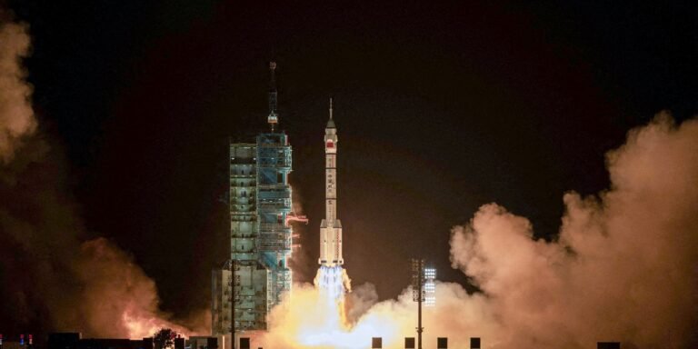 Nighttime rocket launch with a tall rocket lifting off amid orange smoke and a lit launch pad structure.