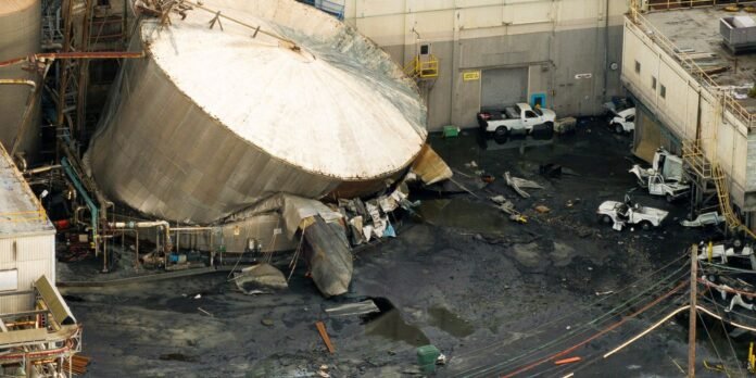 Large cylindrical/ spherical industrial storage tank collapsed in a yard, with debris, puddled oil, and several damaged trucks nearby.