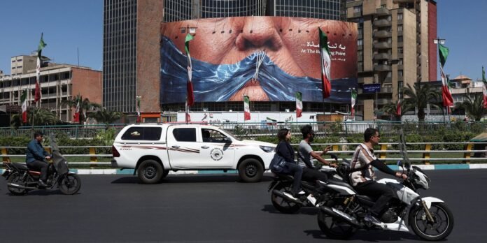 City street scene with a giant billboard showing a masked face, flags along the road, and vehicles including motorcyclists and a white pickup truck.