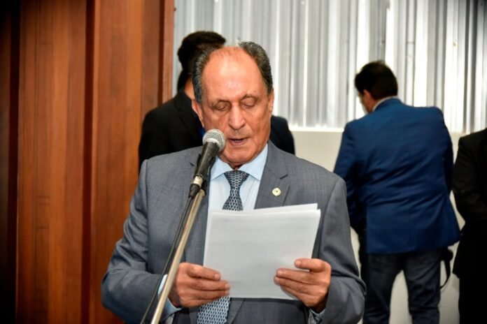 Man in a gray suit reads from papers into a microphone at a formal event.