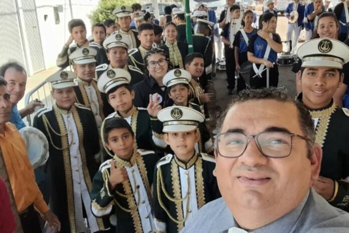 Man wearing glasses taking a selfie with a large group of young band members in green and gold uniforms behind him.