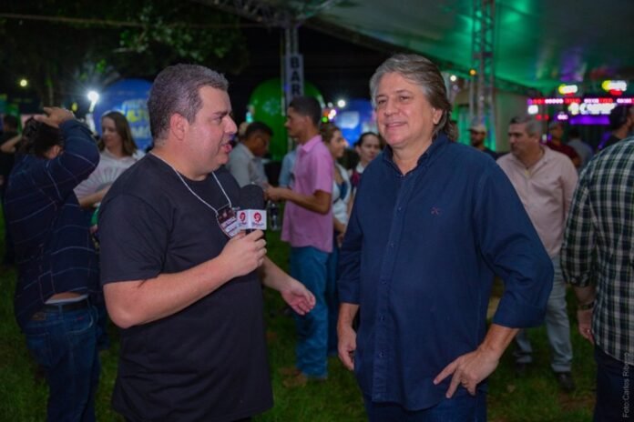 Reporter interviewing a man in a dark shirt at an outdoor evening event with people in the background.