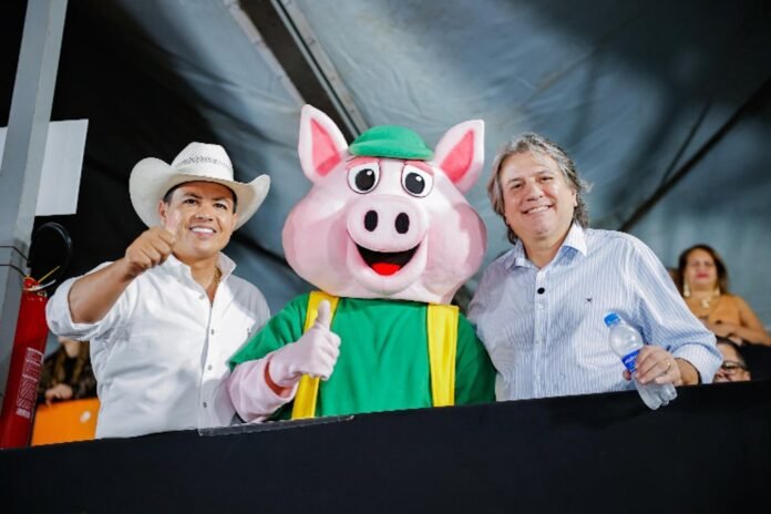 Three people at an event: a man in a white cowboy hat, a pink pig mascot in a green outfit giving a thumbs up, and a man holding a water bottle, all smiling.